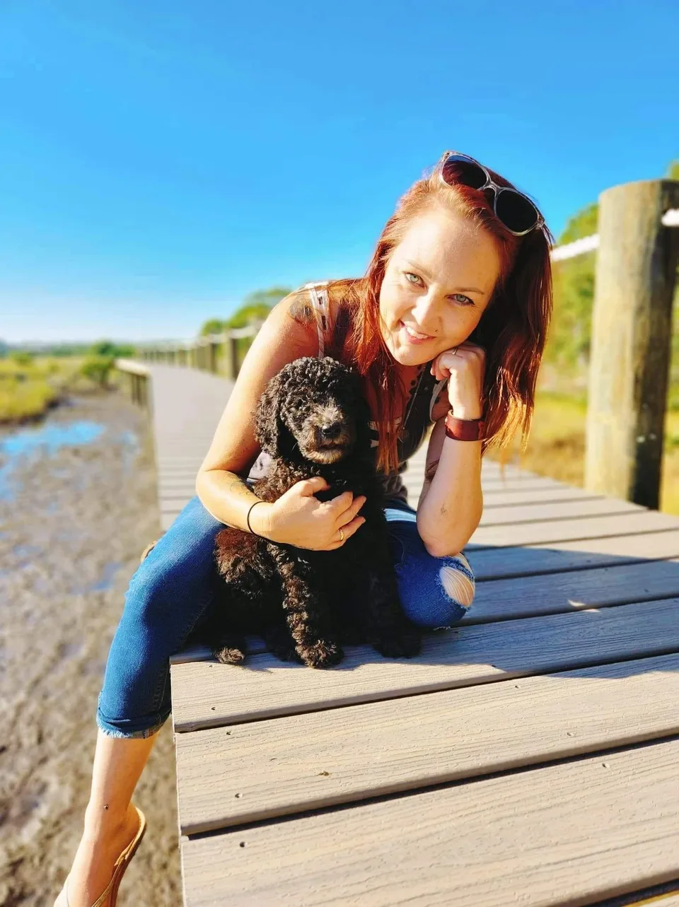 A woman holding a black curly-haired dog beside the water.