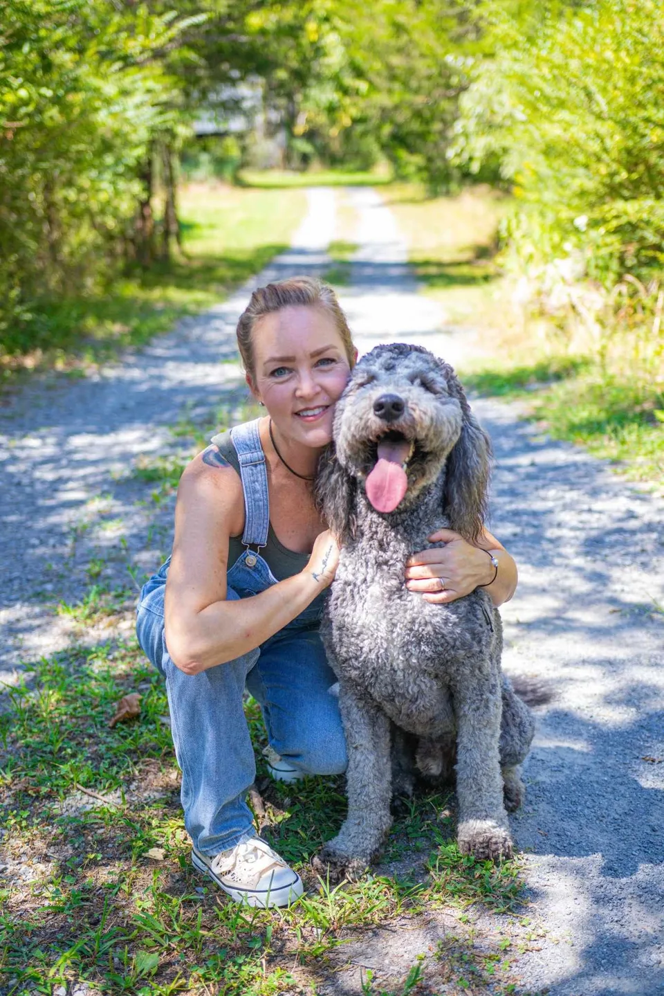 A woman kneeling beside a curly-haired dog on a sunny wooded path.